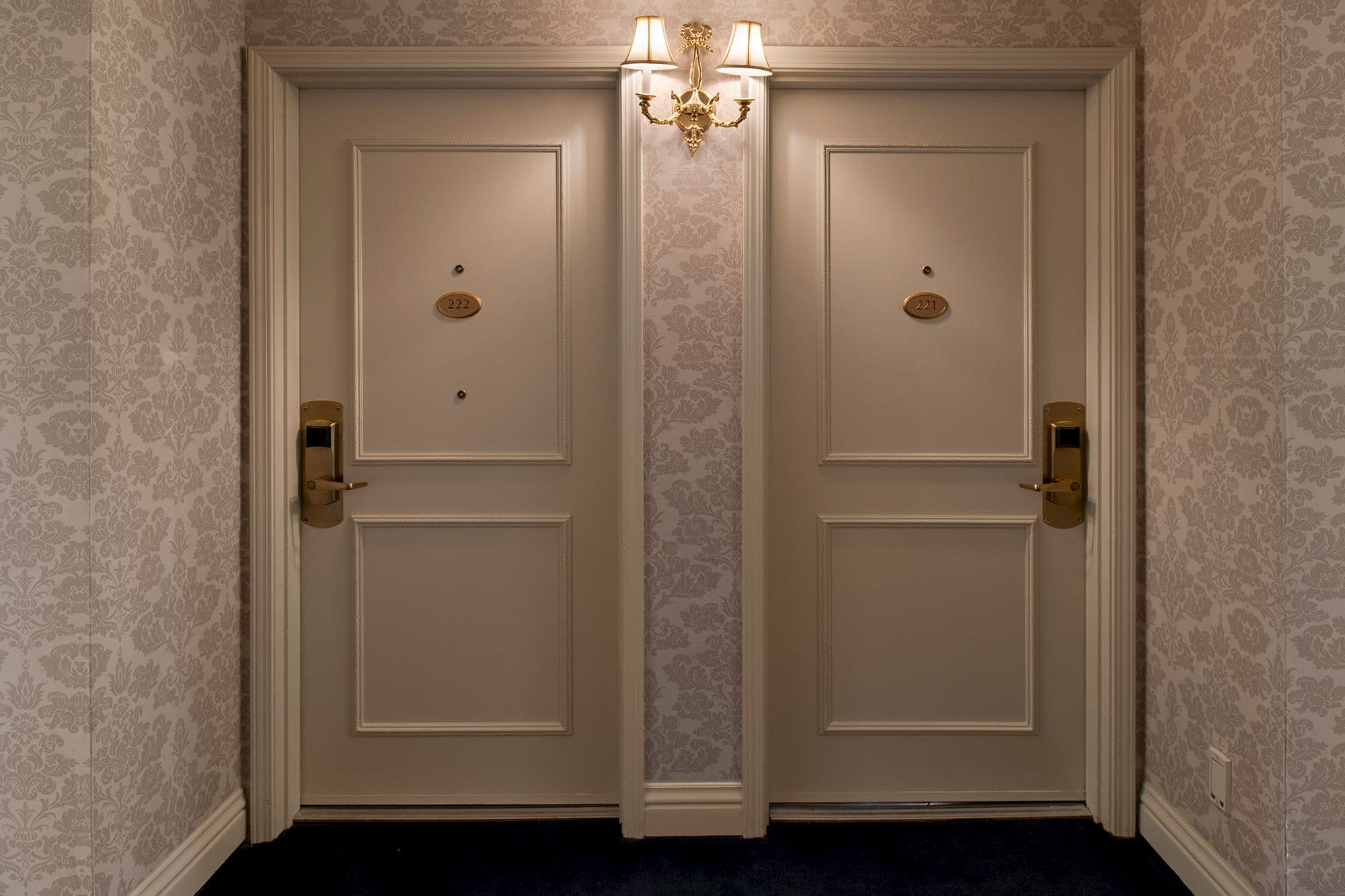 Two paneled guest room doors with brass handles under a brass sconce