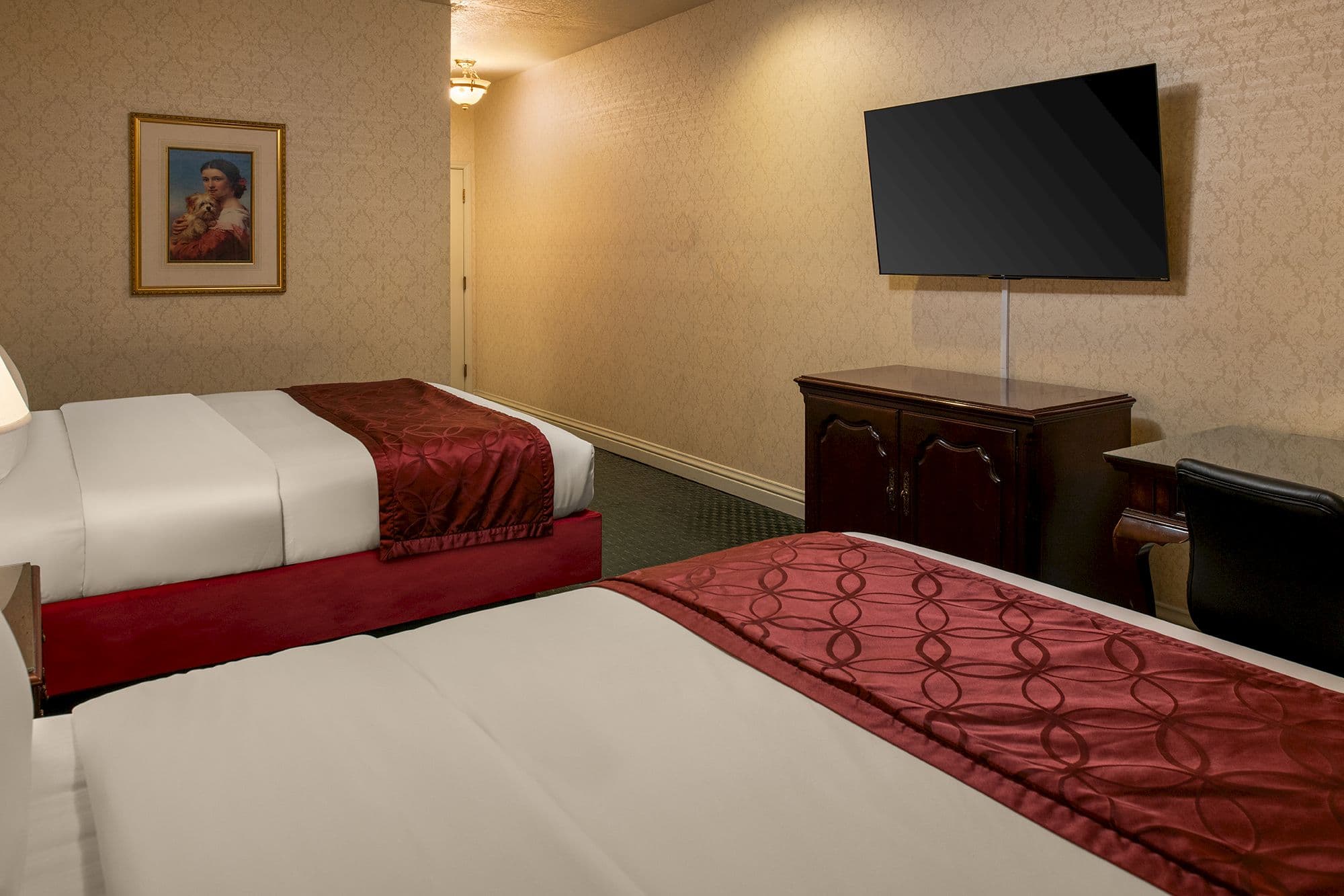 Two queen beds with burgundy runners facing a wall-mounted flat-screen TV and a cherry wood cabinet